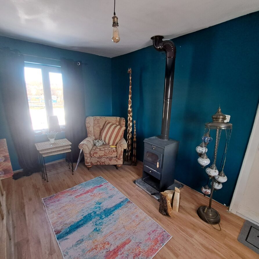 A cosy guest house room at Muddy Puddles Farm features teal walls, a wood-burning stove, a striped armchair, a colourful rug, and tall wooden sticks. Light streams through a window with dark curtains, perfect for a relaxing farm stay.