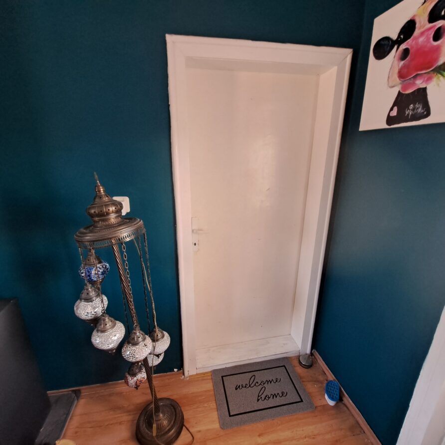 A white door is centred in a teal-walled room at Muddy Puddles Farm guest house. A “welcome home” doormat sits in front. To the left is a decorative stand with hanging ornaments, and to the right, colourful Mickey Mouse artwork brightens the wall.