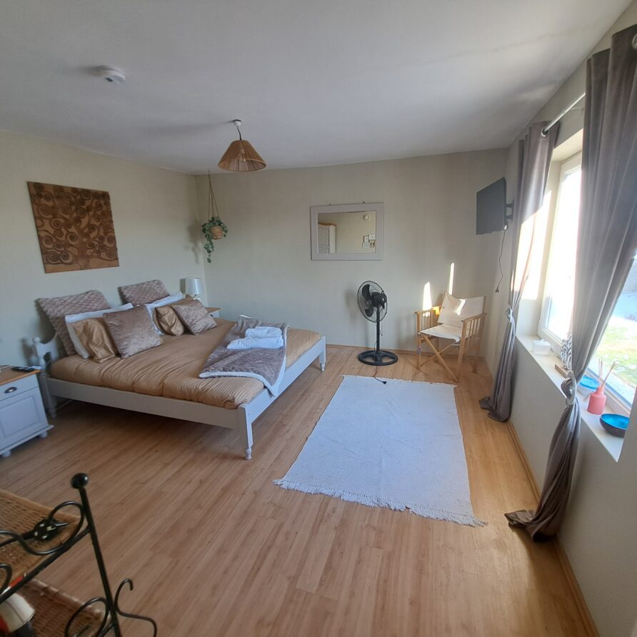 A bright, tidy bedroom at Muddy Puddles Farm with a double bed, beige bedding, two bedside tables, a standing fan, white rug, chair and desk by the window, and wooden flooring. Natural light pours in from large windows with curtains.