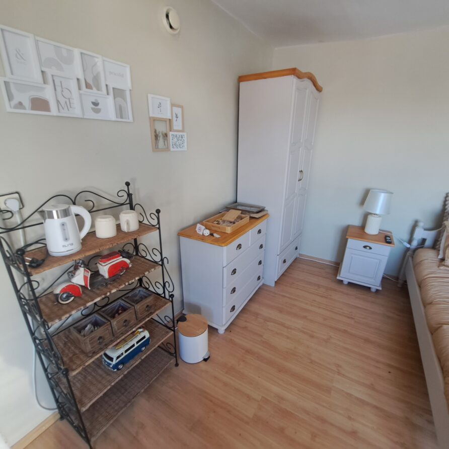 A cosy bedroom at Muddy Puddles Farm with light-coloured walls and wooden flooring, featuring a bed with beige bedding, a white wardrobe, a small cupboard, a lamp, and a metal shelf holding a kettle, cups, and decorative items.