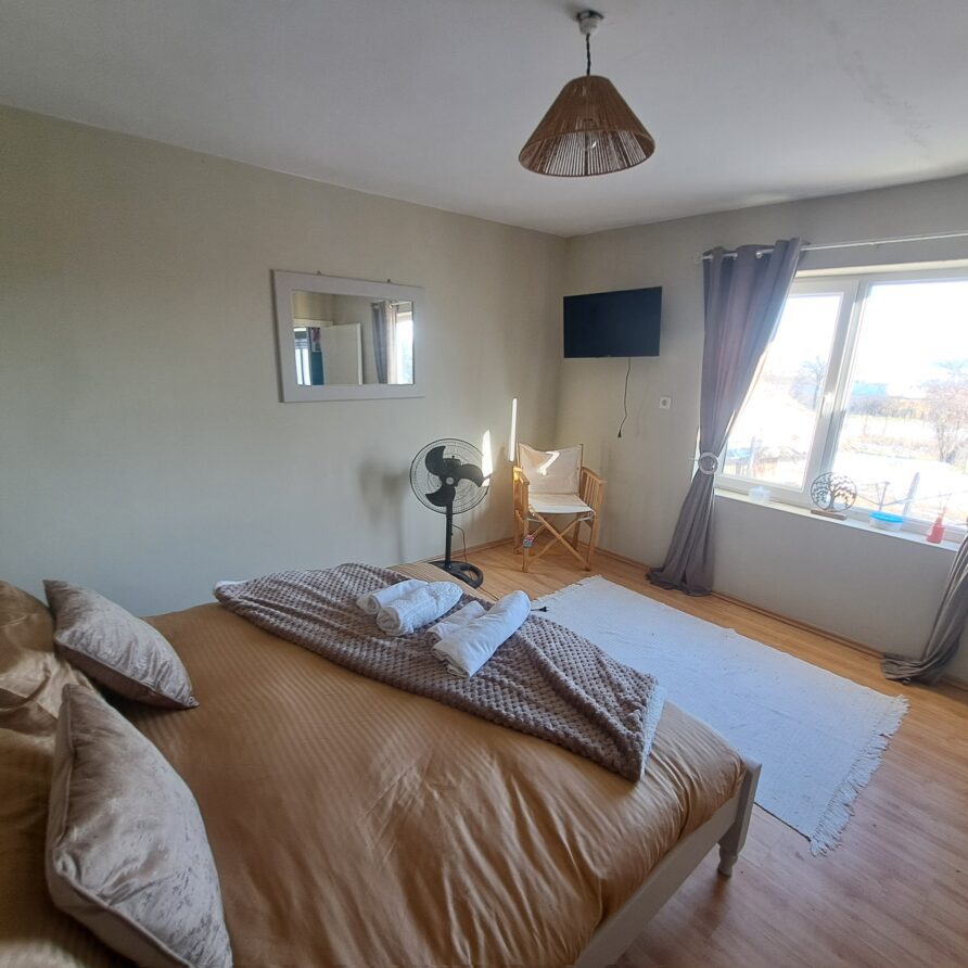 A bright guest house bedroom with a double bed, tan bedding, and pillows. There’s a window with grey curtains, a wall-mounted TV, standing fan, chair, white rug, hanging plant, and mirror—perfect for a cosy farm stay at Muddy Puddles Farm.