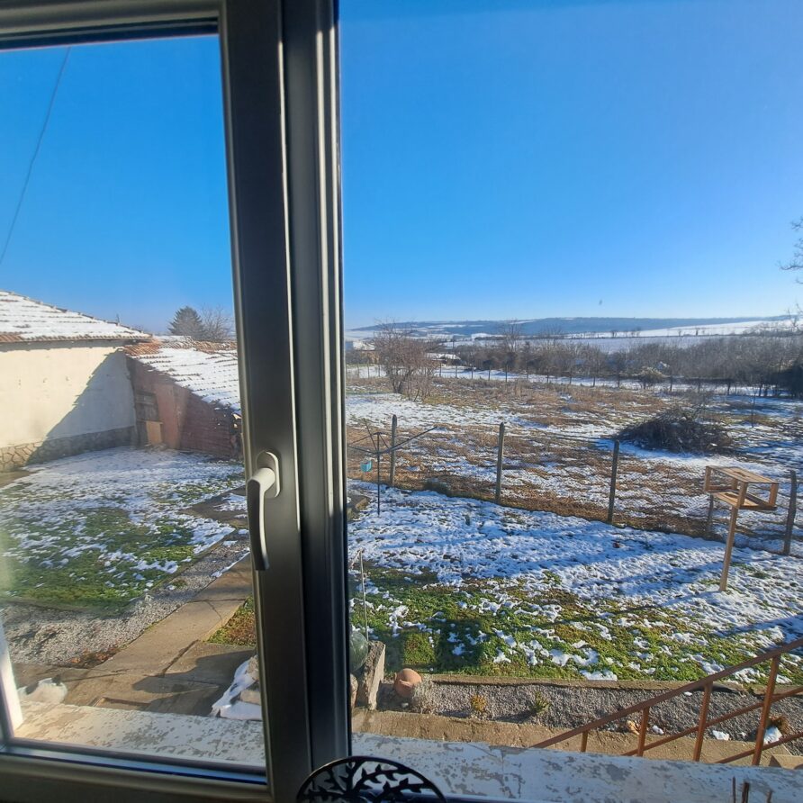 View through a window showing a snowy back garden with patches of grass, a small white guest house on the left, bare trees, and a clear blue sky on a bright winter day—perfect for a peaceful Kovachevets farm stay.
