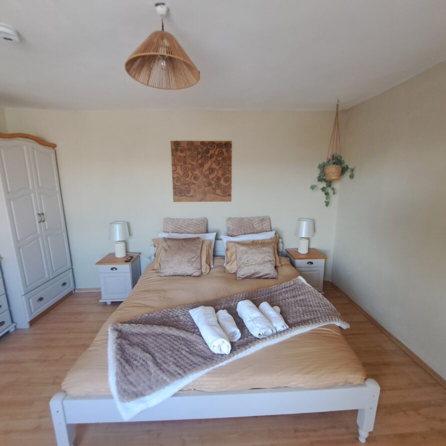 A neatly made double bed with beige bedlinen and rolled towels, flanked by two bedside tables with lamps. This cosy, light-filled guest house bedroom in Kovachevets features a wardrobe, chest of drawers, hanging plant, and wall art.