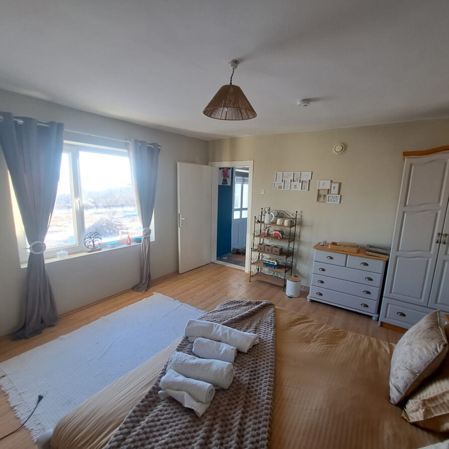 A cosy bedroom at Muddy Puddles Farm guest house in Kovachevets, with a large bed, folded towels, beige bedding, a white wardrobe, chest of drawers, wall shelves, and natural light through grey-curtained windows. Door leads to another room.