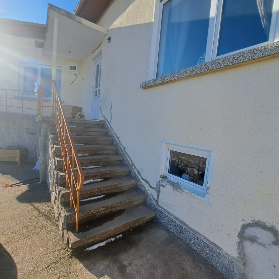 A concrete staircase with a metal railing leads to a white door on a beige house at Muddy Puddles Farm. Large windows reflect the sky whilst two dogs relax near the steps on this sunny farm stay in Kovachevets.