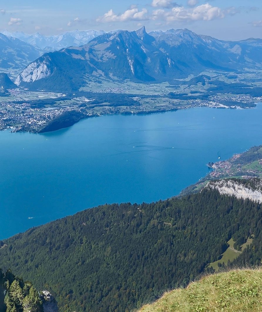 A scenic view of a deep blue lake surrounded by green hills and forested mountains, with charming guesthouses in small towns along the shoreline and tall snow-capped peaks in the background under a partly cloudy sky.