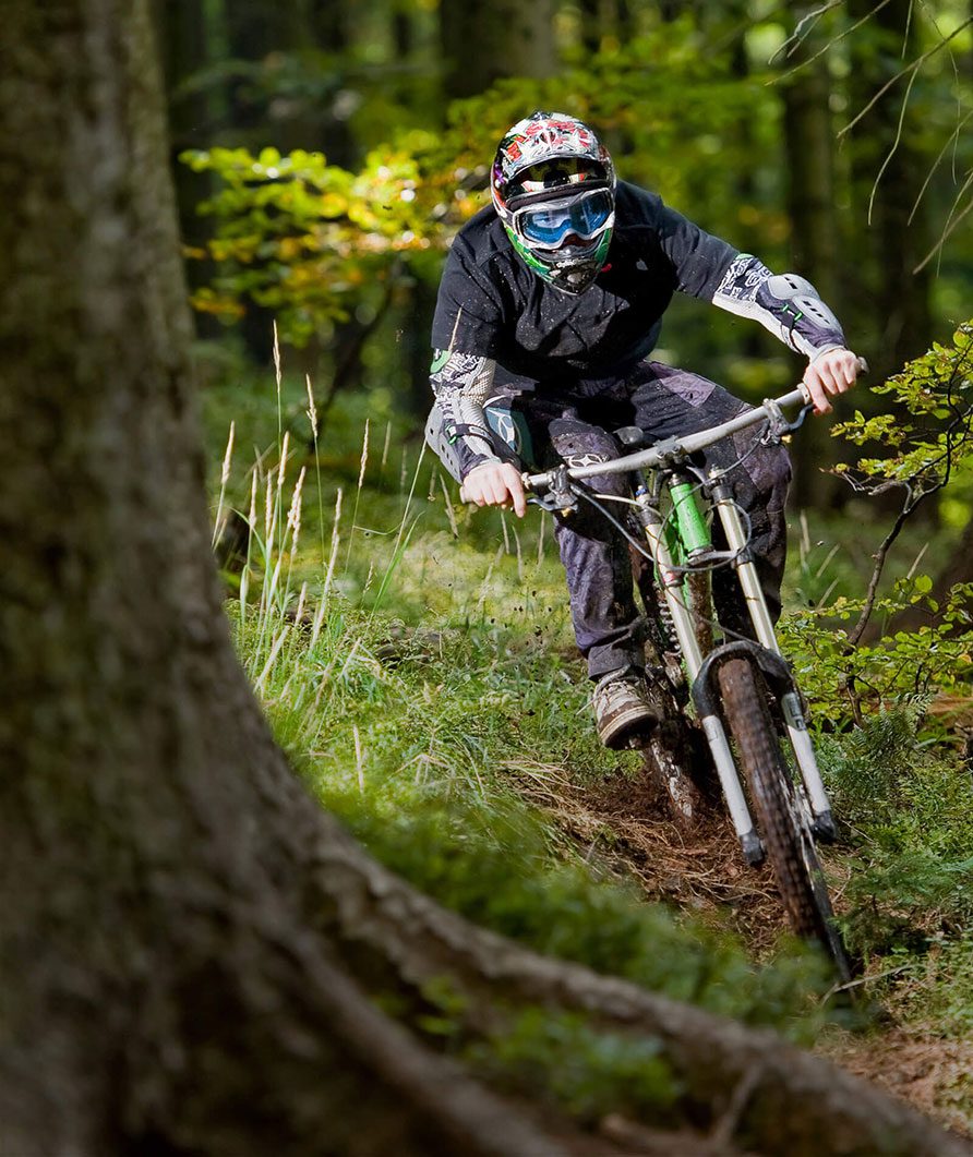 A mountain biker wearing protective gear and a helmet rides quickly down a narrow dirt track at Muddy Puddles Farm in Kovachevets, steering around a large tree near the guest house through the lush, green forest.