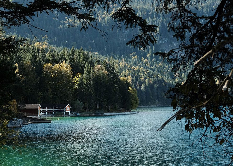 A serene lake with turquoise water is bordered by dense evergreen trees. Near the shore, a small white and red boathouse and a cosy guest house sit under the forest’s shade, with mountains in the background.