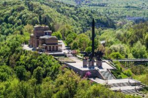 Aerial view of a large monument with a tall obelisk surrounded by statues, set on a raised platform amid lush green trees, with a guest house to the left and a forested landscape in the background.