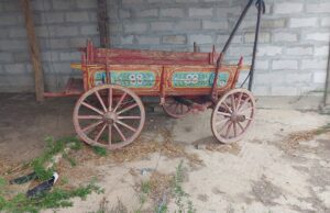 A colourful, old-fashioned wooden cart with hand-painted floral designs stands on a dirt floor near a stone wall at a charming Kovachevets guest house. The cart, with large spoked wheels, sits under a simple shelter with wooden posts.