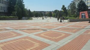 A large open plaza with red and beige chequered tiles, lined with trees and buildings on both sides. Several people are walking or standing around, creating a lively scene reminiscent of a bustling guest house under a clear and sunny sky.