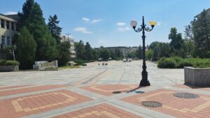 A spacious, paved square with a decorative black lamp post in the foreground, surrounded by trees and buildings under a clear blue sky. Nearby, Muddy Puddles Farm offers a peaceful farm stay experience in charming Kovachevets. A few people walk in the distance.