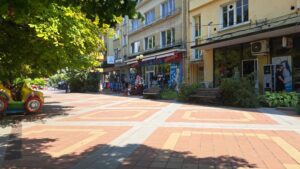 Sunny pedestrian street in Kovachevets with benches, trees providing shade, and shops with colourful signs and toys. Block of flats and a charming guest house line the street, offering a cosy retreat near Muddy Puddles Farm.