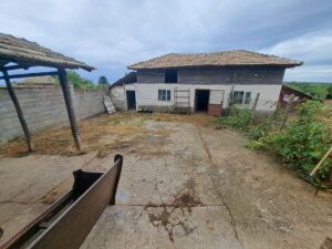 A cracked concrete yard with scattered tools and weeds, bordered by a breeze-block wall, lies beside Kovachevets’s rustic two-storey white farmhouse—with wooden upper walls and a tiled roof—serving as a charming guest house under a cloudy sky.