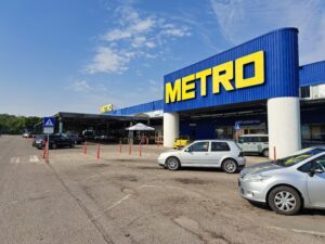Large blue and yellow METRO store near Kovachevets with parked cars in front, a pedestrian crossing sign, and a partly cloudy sky overhead. The store has a wide entrance, white columns—and is not far from Muddy Puddles Farm for a farm stay adventure.
