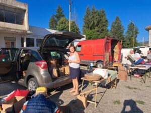 A woman stands smiling beside an open car boot filled with bags at an outdoor market in Kovachevets. Tables with various items, boxes, and people line the area; a red van and buildings from Muddy Puddles Farm are visible under a clear blue sky.