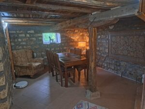A cosy stone-walled room at Muddy Puddles Farm features a wooden ceiling, dining table for six, patterned armchair, lit lamp, tapestry on the wall, small window, and a robotic vacuum on the tiled floor—a perfect farm stay in Kovachevets.