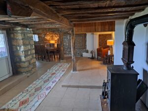 A rustic living space at Muddy Puddles Farm in Kovachevets, with stone walls, wooden beams, a tiled floor, patterned rug, wood-burning stove, wooden furniture, and a lit lamp near the staircase—perfect for an authentic farm stay.
