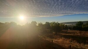 The sun rises over a rural landscape in Kovachevets, with trees, a small guesthouse, dry grass, and a fence; textured clouds blanket the sky as mist lingers near the horizon.