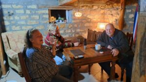 Three adults sit around a wooden table in a cosy guest house in Kovachevets. Two women and a man enjoy drinks and conversation under warm lighting, surrounded by rustic stone walls. A lamp and small objects decorate the farm stay setting.