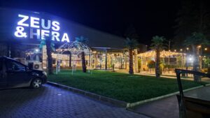 Night-time view of a brightly lit restaurant named ZEUS & HERA, featuring illuminated outdoor seating areas with fairy lights, artificial palm trees, glass domes, and a parked black car in front—perfect for farm stay guests from Muddy Puddles Farm or nearby guest houses.