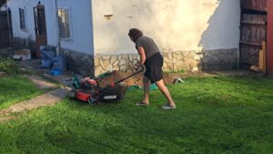 A person in a green T-shirt and black skirt is pushing a lawnmower on the grassy garden of a guest house with a stone foundation in Kovachevets on a sunny day.