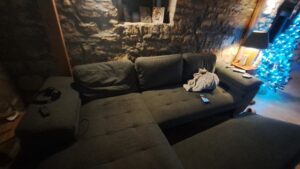 A dimly lit lounge in the guest house at Muddy Puddles Farm, Kovachevets, features a dark corner sofa with a towel and mobile on the seat, headphones and remote on the armrest, a stone wall, lit lamp, and blue-lit Christmas tree.