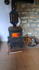 A small brown dog lies on a blanket in a pet bed next to a lit wood-burning stove in a guest house room with stone walls and a tiled floor, offering cosy comfort during your farm stay in Kovachevets.
