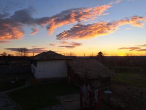 A rural guest house with a tiled roof stands under a colourful sunset sky with dramatic orange clouds. Washing hangs on a line in the garden, and a dog roams the grassy area near the fence at Muddy Puddles Farm.