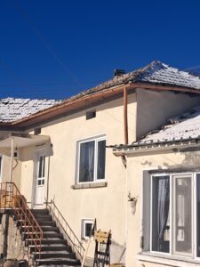 A beige house at Muddy Puddles Farm with a snow-covered roof, white-framed windows, and a staircase with an orange handrail leading to the entrance under a clear blue sky offers a cosy farm stay.