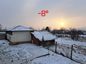 A snowy rural scene in Kovachevets shows two houses and a fenced garden at sunrise or sunset. The -8°C temperature glows red atop the image as snow blankets the ground and rooftops—perfect for a cosy farm stay at Muddy Puddles Farm.