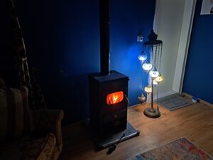 A wood-burning stove with a glowing fire sits next to a standard lamp with colourful glass lanterns, against a dark blue wall in a cosy Muddy Puddles Farm stay room with a patterned rug and armchair.