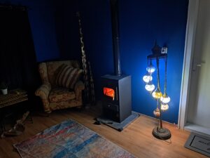 A cosy living room at Muddy Puddles Farm features a patterned armchair, a wood-burning stove with a fire inside, a mosaic floor lamp, small table, and blue wall—perfect for relaxing after exploring the Kovachevets farm stay.