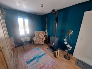 A cosy guest house room in Kovachevets with teal walls, a wood-burning stove, patterned armchair, side table, tall giraffe sculptures, colourful rug, wooden floor, and a window with black curtains letting in sunlight—a perfect farm stay escape.