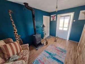 A cosy guest house room at Muddy Puddles Farm with teal walls, a striped sofa, wood-burning stove, giraffe sculptures, colourful rug, light wood floors, cow artwork on the wall, and sunlight streaming through a glass door.