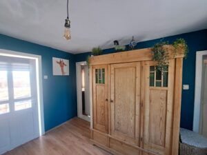 A bright guest house room at Muddy Puddles Farm with blue walls, wooden floor, and a large wooden wardrobe decorated with plants and animal figurines. A giraffe picture hangs on the wall as sunlight streams through the window.