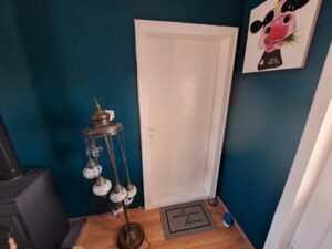 A white door in a teal-painted room at Muddy Puddles Farm, with a welcome home doormat, a decorative stand with hanging ornaments, and wall art featuring a colourful, abstract animal face—perfect for your cosy farm stay.