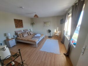 A cosy, well-lit bedroom in a guest house in Kovachevets, featuring a large bed with beige bedding, white bedside tables, a fan, a white chair and desk, wall mirror, wooden floor, and large windows with brown curtains.