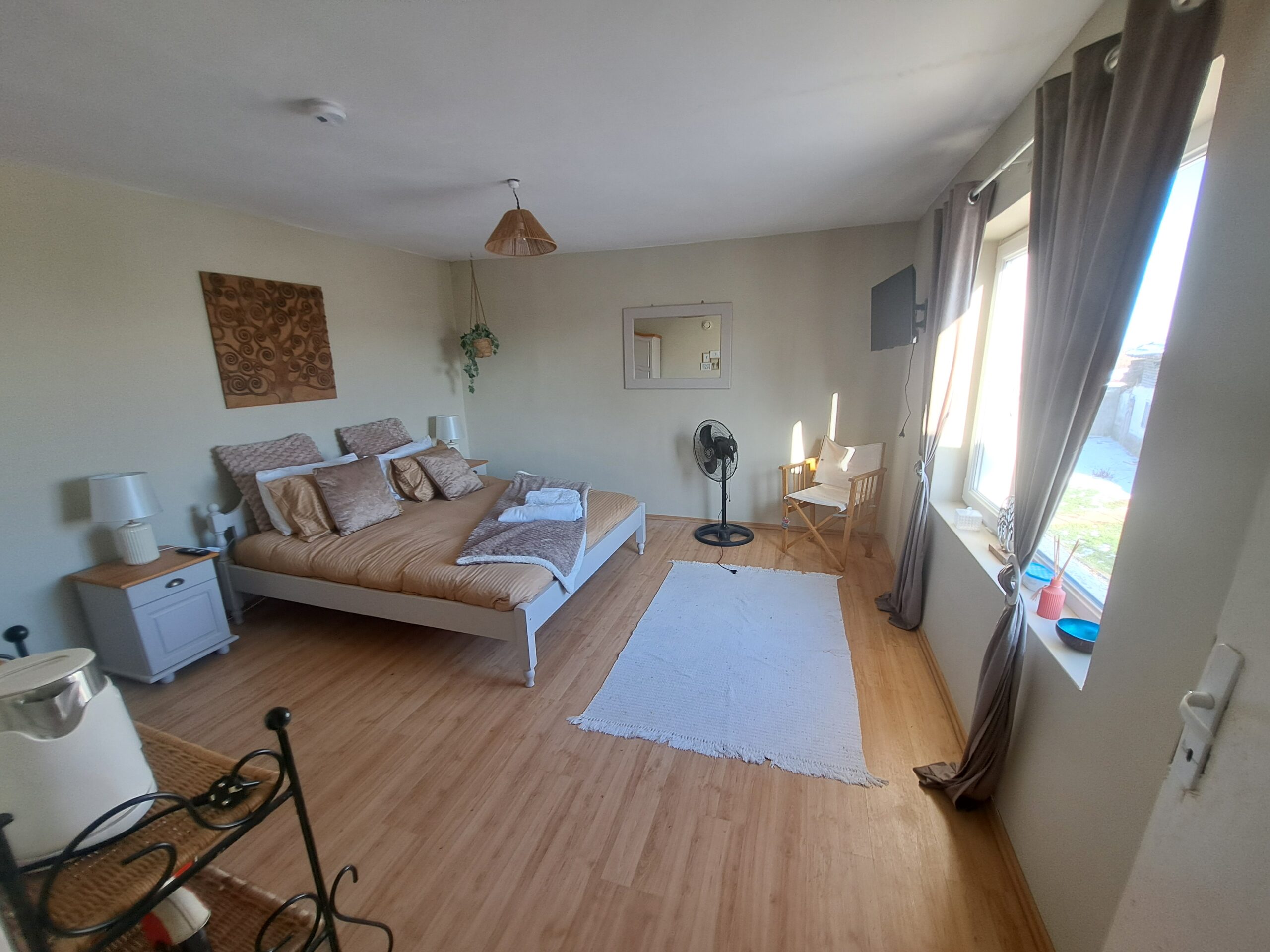 A bright, cosy bedroom at the Kovachevets guest house features a large bed, bedside tables, a white rug, fan, wooden chair, and large windows with brown curtains letting in natural light over light wood floors and soft-coloured walls.