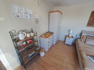 A cosy bedroom at Muddy Puddles Farm in Kovachevets features light wood flooring, beige bedding, a white bedside table and lamp, a wardrobe, chest of drawers, black metal shelf with kitchen items, and charming wall art.