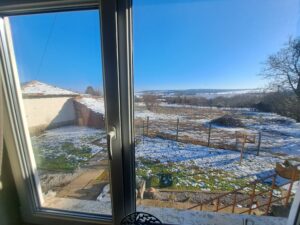 A view through a window at a Kovachevets farm stay reveals a sunlit yard with patches of melting snow, a small tiled-roof guest house on the left, bare trees, and open fields stretching under a clear blue sky in the distance.