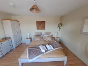 A cosy bedroom in Muddy Puddles Farm guest house, Kovachevets, featuring a double bed with beige bedding, neatly folded towels, two bedside tables with lamps, a wardrobe, chest of drawers, wall art, and a charming hanging plant.