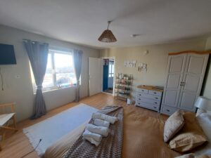 A cosy, sunlit bedroom in the guest house at Kovachevets, with a neatly made bed, towels on top, light-coloured curtains, a large window, chest of drawers, wardrobe, and an open door revealing Muddy Puddles Farm next door. Décor includes wall art and a woven lampshade.