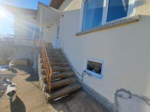 A set of concrete steps with a metal handrail leads to a white door on a cream-coloured guest house. Two dogs stand on the sunlit ground near the steps, and a window is visible on the right, hinting at relaxed moments during your Kovachevets farm stay.