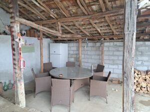 A rustic outdoor seating area at Muddy Puddles Farm features a round glass table and six wicker chairs under a wooden, tiled-roofed pergola. A fridge, stacked firewood, and hanging ropes complete this cosy farm stay setting.
