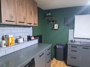 Modern kitchen at Muddy Puddles Farm with grey worktops and cupboards, light grey metro tile splashback, wooden wall units, green wall, shelves with plants and decor, rubbish bin, and a small appliance—your cosy Kovachevets farm stay awaits.