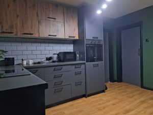 Modern kitchen at Muddy Puddles Farm with grey lower cabinets and wooden uppers, white subway tile splashback, built-in oven, microwave, countertop appliances, green walls, and wood-style flooring under ceiling lights—perfect for your farm stay in Kovachevets.