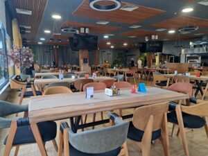 Spacious modern restaurant with wooden tables and colourful cushioned chairs, located near Kovachevets. The well-lit room features large windows, plants, and a few people seated in the background—perfect for guests enjoying a farm stay.
