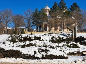 A snow-covered park in Kovachevets with evergreen shrubs, a stone path, and a historic domed guest house partially hidden by leafless trees under a clear blue sky.