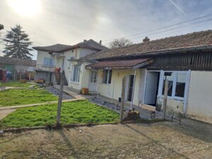 A weathered, single-storey guest house with peeling paint and a tiled roof stands in a garden with green grass and clothes hanging on a line; sunlight shines through a partly cloudy sky in Kovachevets.